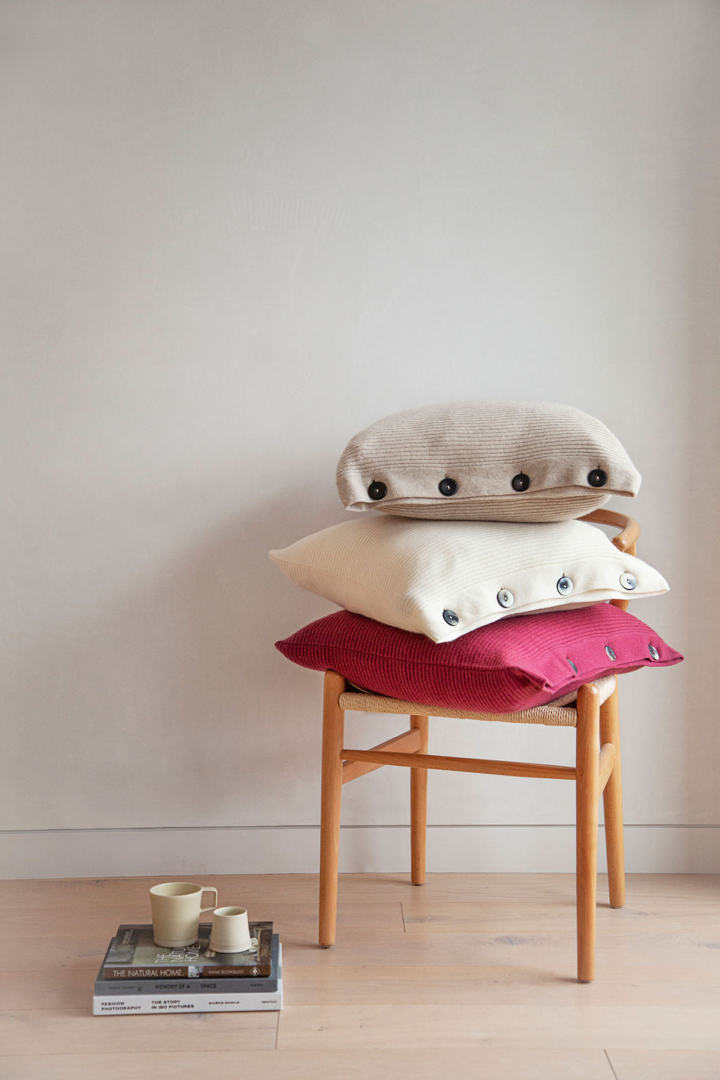 Pictured: latte, white, and poppy pillows stacked on a chair. On the floor next to the chair is a mug and cream holder on top of three books.